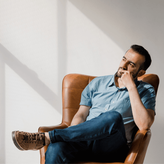 man sitting in a leather brown chair, thinking position with his left hand on his chin and his left leg crossed over his right knee.