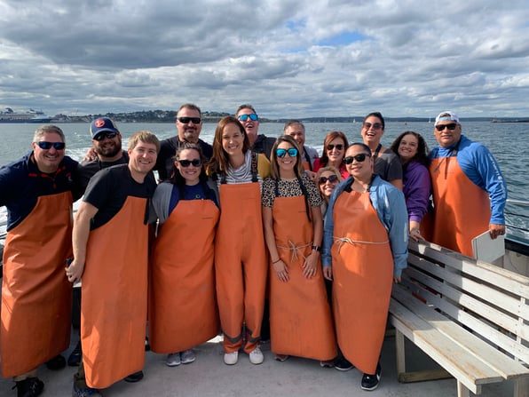 A group of Compass Award winners wearing orange fishing aprons pose on a boat deck, with a harbor, city skyline, and cruise ship behind them.