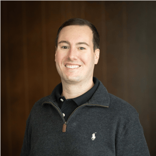 Headshot of Ethan Long smiling, wearing a dark gray quarter-zip sweater over a navy collared shirt, against a dark wood background.