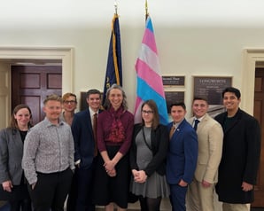 A group of MMAA members is seen smiling in front of the trans flag and a military flag.