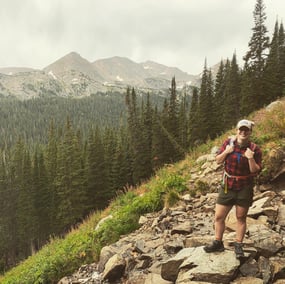 Remy stands on the side of a rocky mountain with a backpack on, and pine trees/the skyline behind her.