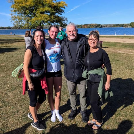 JoAnna and Renee smile in a group shot outdoors by a lake at a cross country meet.