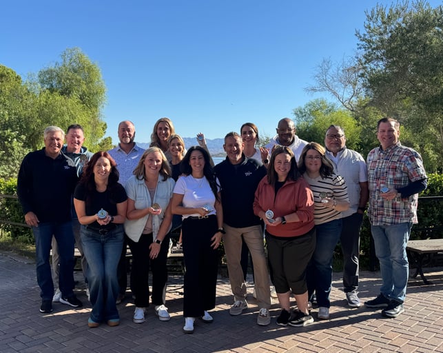 Compass Award winners stand together on a sunny brick patio, holding up their compass awards, with trees, a lake, and distant mountains under a clear blue sky behind them.