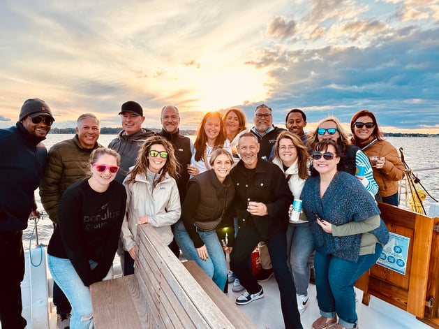 The Compass crew gathers on the deck of a sailboat at sunset, smiling with drinks in hand as the sun breaks through the clouds over the water.