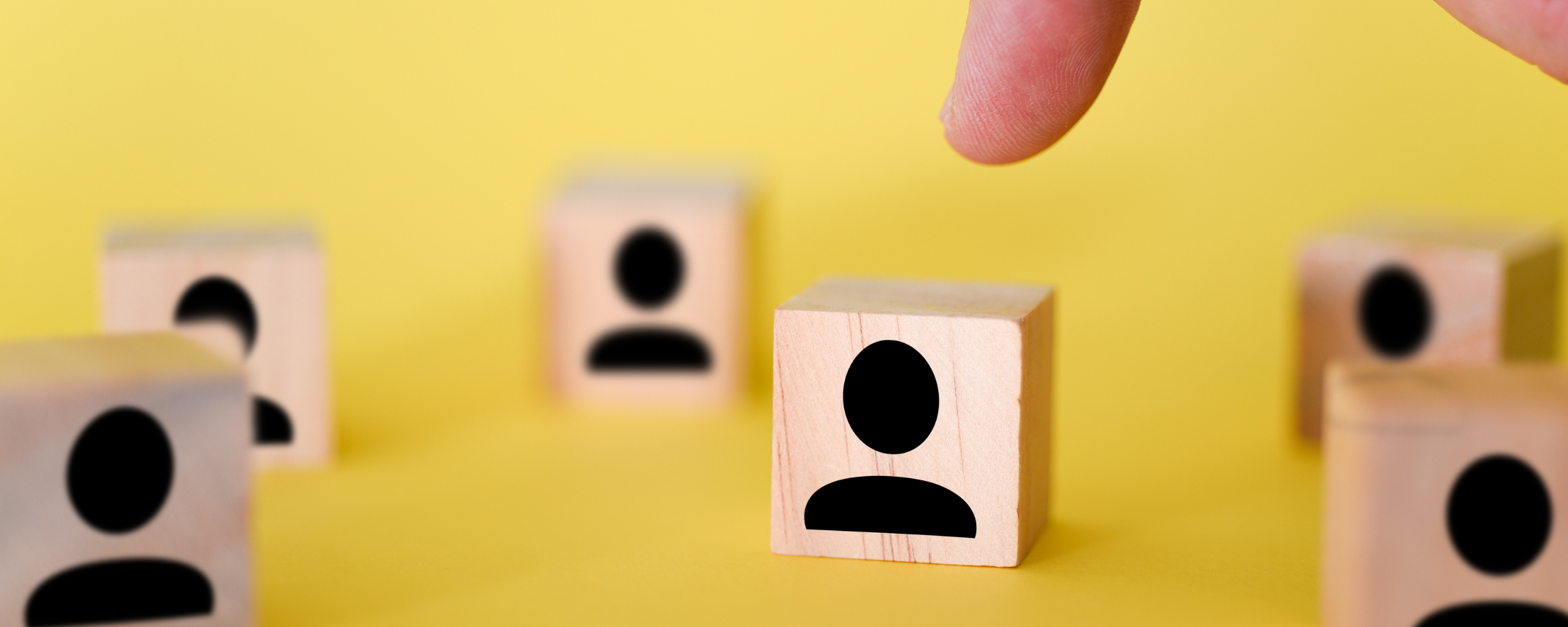 Wooden blocks representing people arranged on a yellow background, with a hand moving one of the blocks into place, symbolizing hiring and workforce planning.