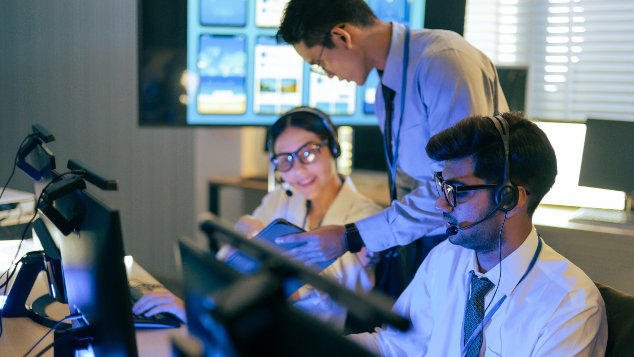A workplace photo showing three professionals wearing headsets in a modern office environment. One person is seated at a computer in the foreground, another is seated behind him smiling, and a third person stands between them pointing at a phone screen while collaborating.