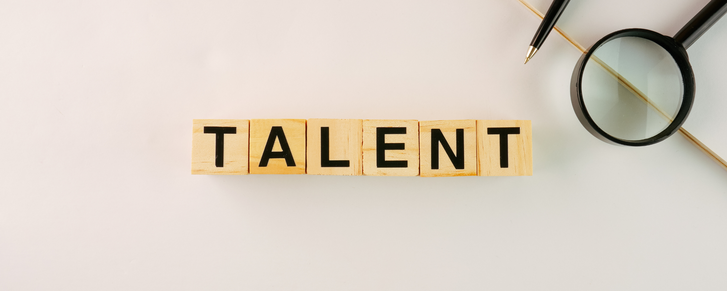 Wooden blocks spelling “TALENT” arranged on a desk beside a magnifying glass and pen, representing talent strategy and workforce planning.